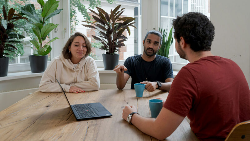 Three people at a table having a discussion, one man points at a laptop